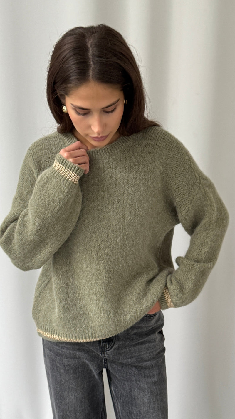 Woman wearing an olive green knitted sweater with beige trim detail on the cuffs and hem, paired with dark grey denim jeans