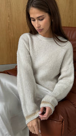 Woman sitting on a brown leather chair, wearing a cream-colored knit sweater with subtle gold detailing on the cuffs, paired with a silky white skirt